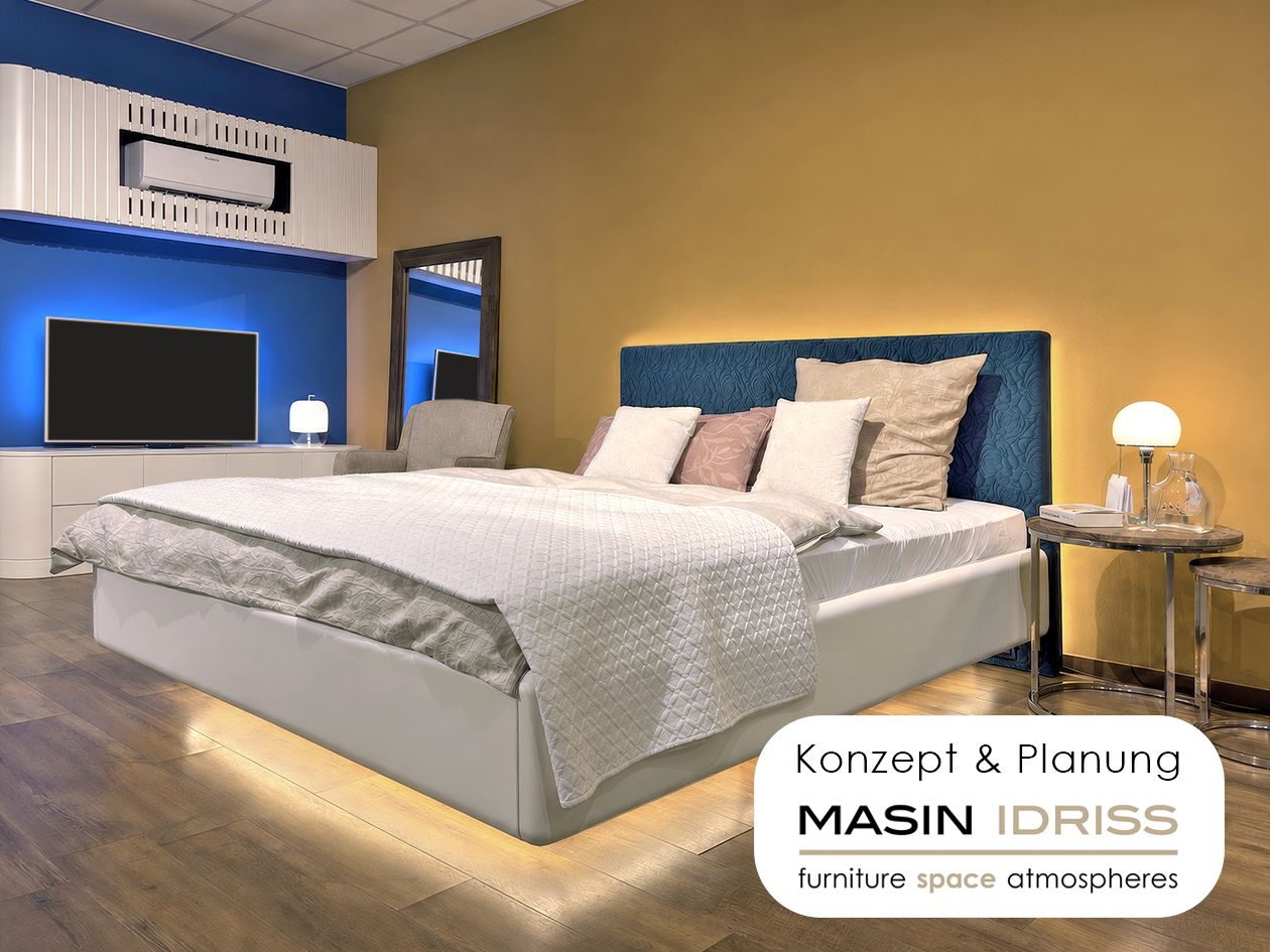 Musterzimmer von Goethe 64: Doppelbett mit beleuchtetem Sockel vor ockerfarbener Wand, Plakette „Konzept & Planung – Masin Idriss“
